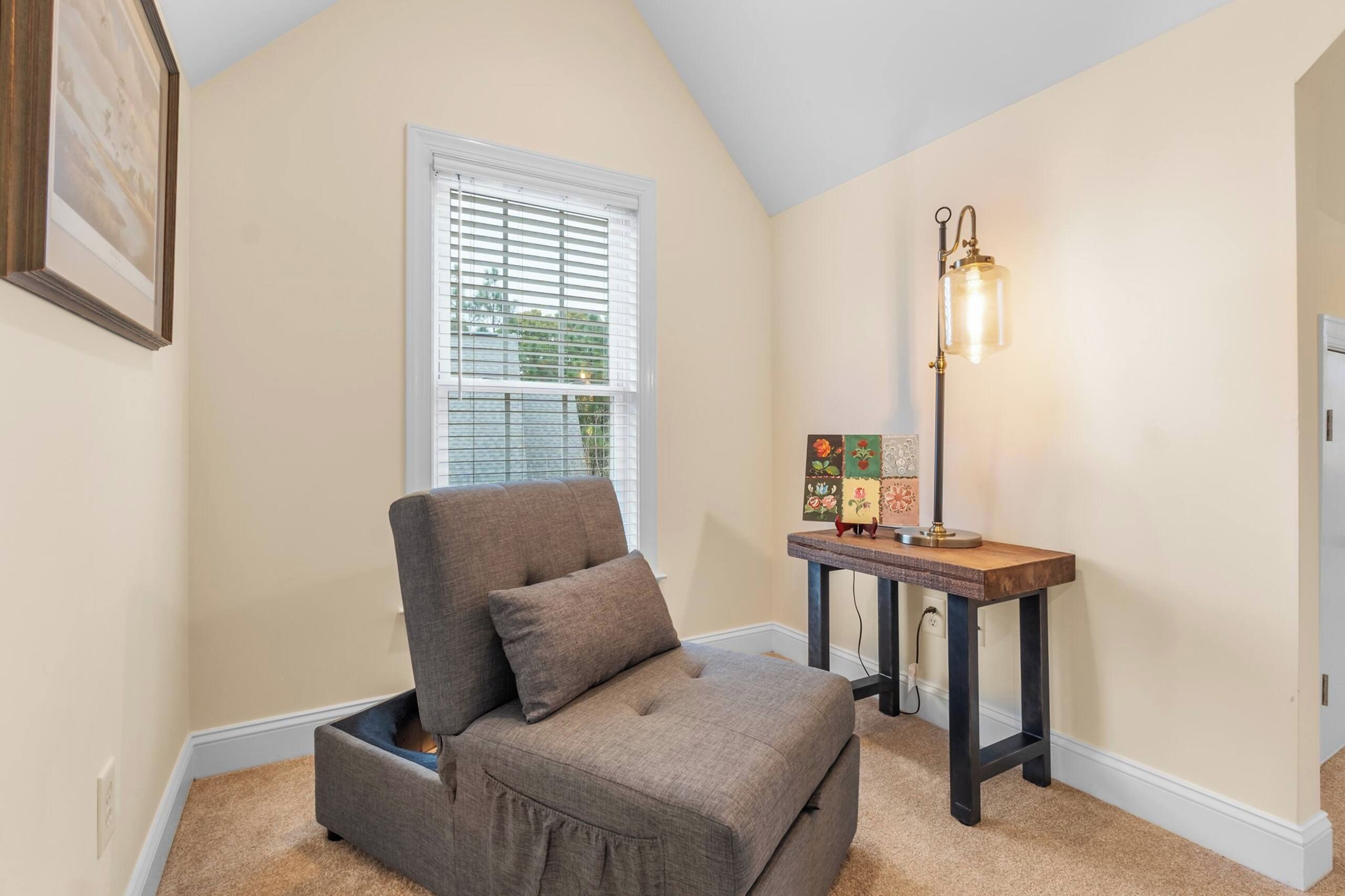 A room with a gray upholstered chair, a small table with a lamp, and a window with blinds.
