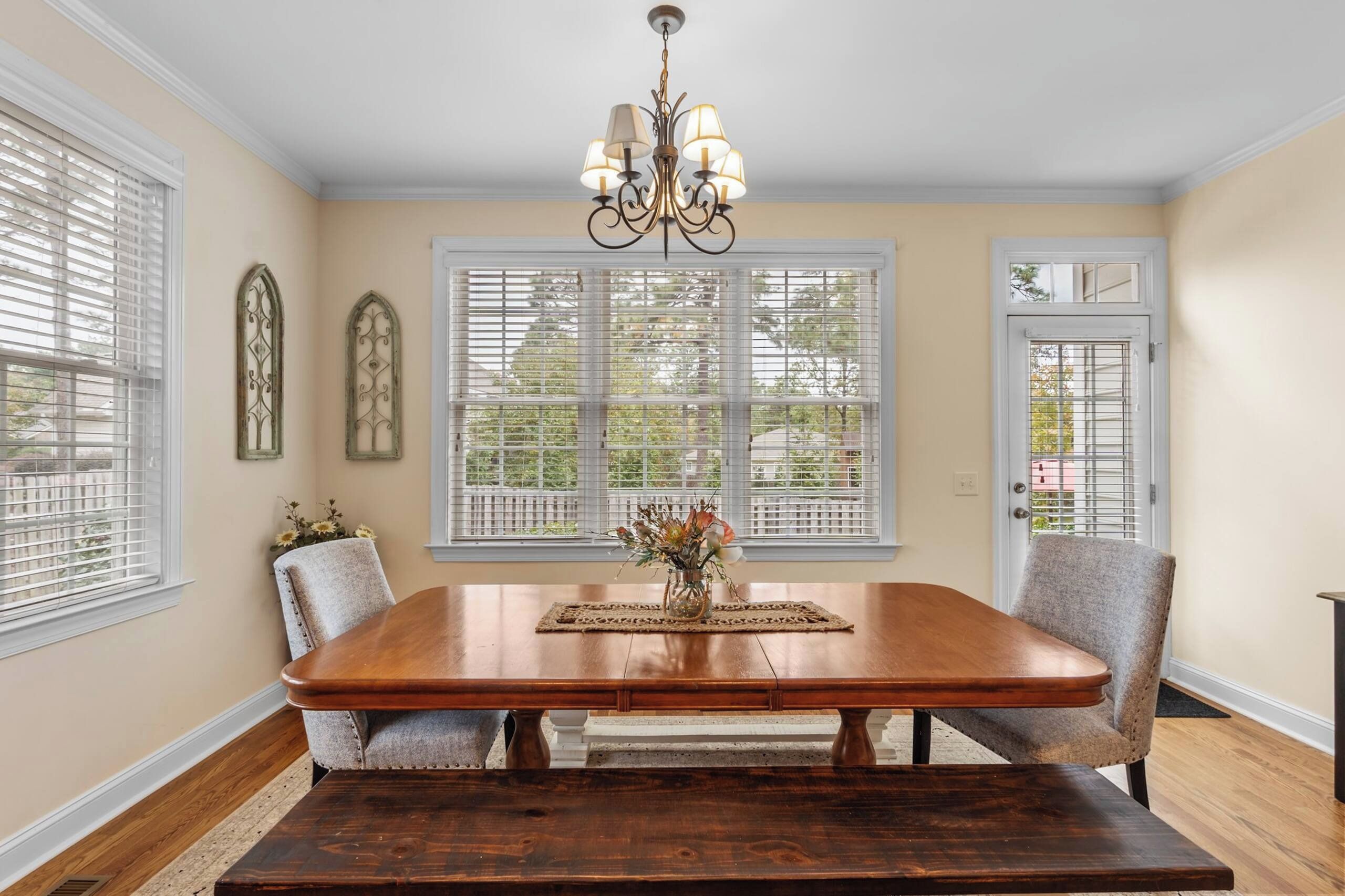 A dining room with a wooden table, two chairs, a chandelier, and a window with blinds.