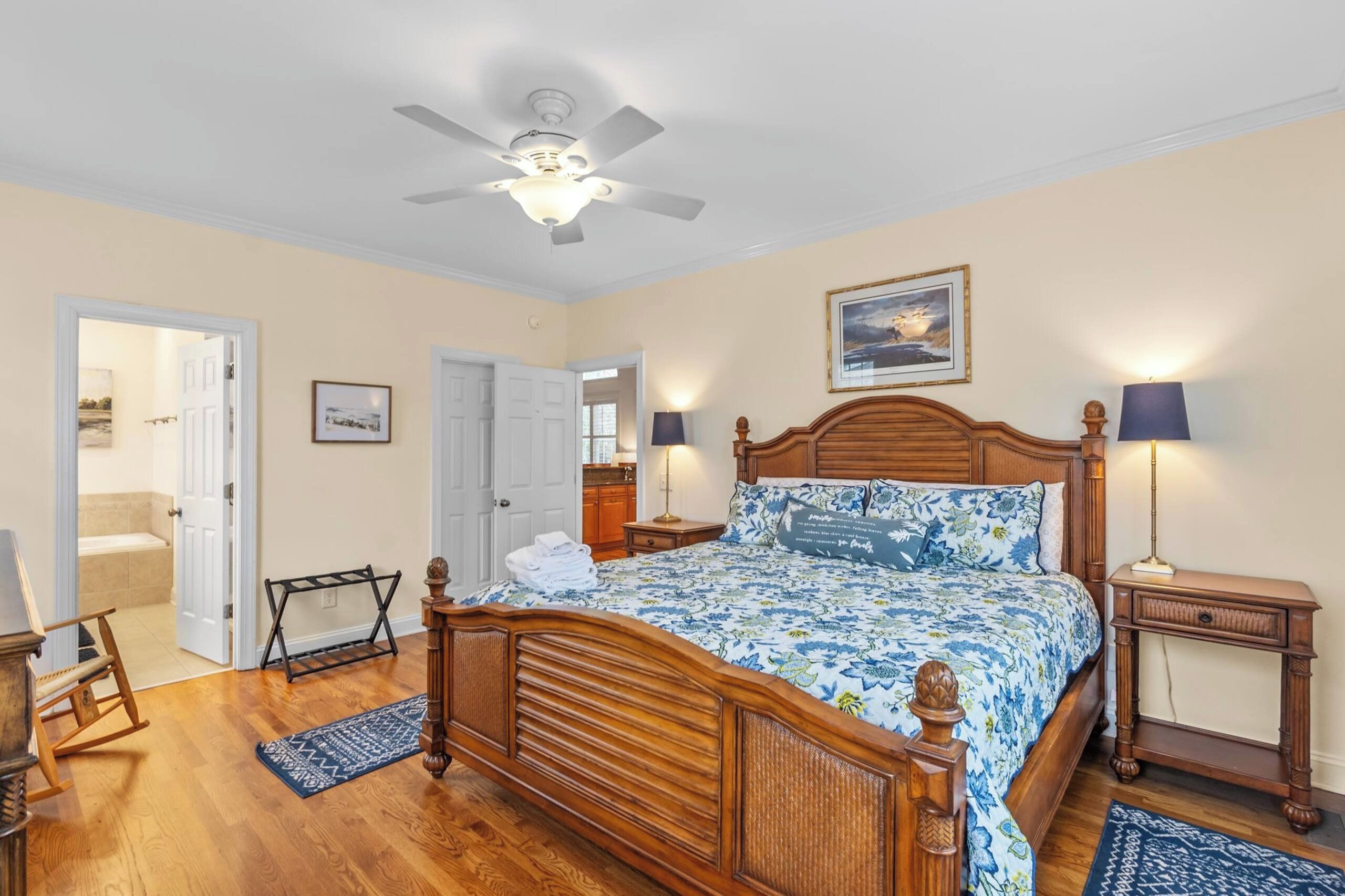A bedroom with a wooden bed, a ceiling fan, a nightstand, a lamp, a rocking chair, and a bathroom visible through an open door.