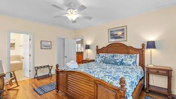 A bedroom with a wooden bed, a ceiling fan, a nightstand, a lamp, a rocking chair, and a bathroom visible through an open door.
