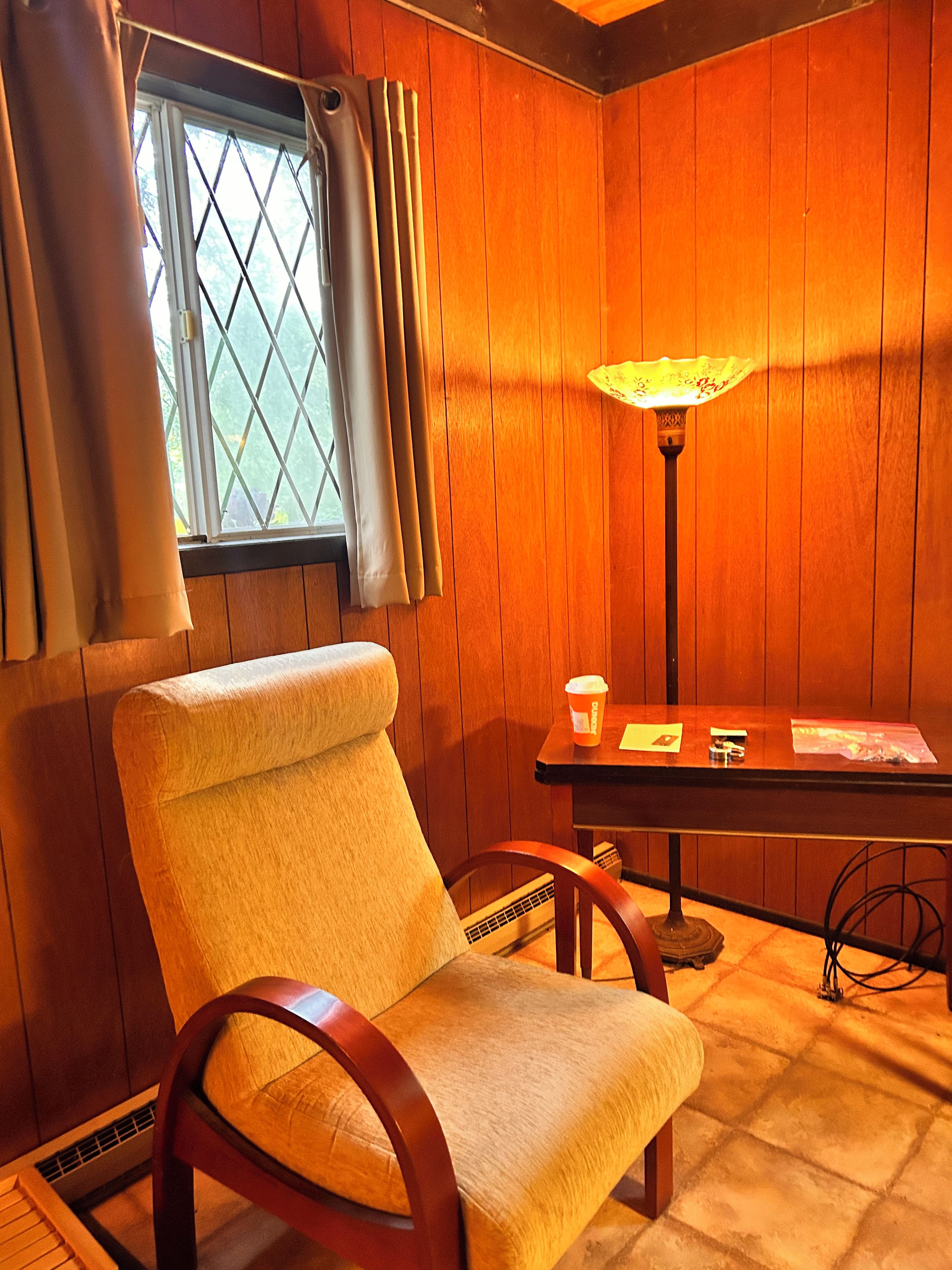 A room with wooden paneling, a window with curtains, a chair, a table with a lamp, and a cup.