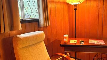 A room with wooden paneling, a window with curtains, a chair, a table with a lamp, and a cup.