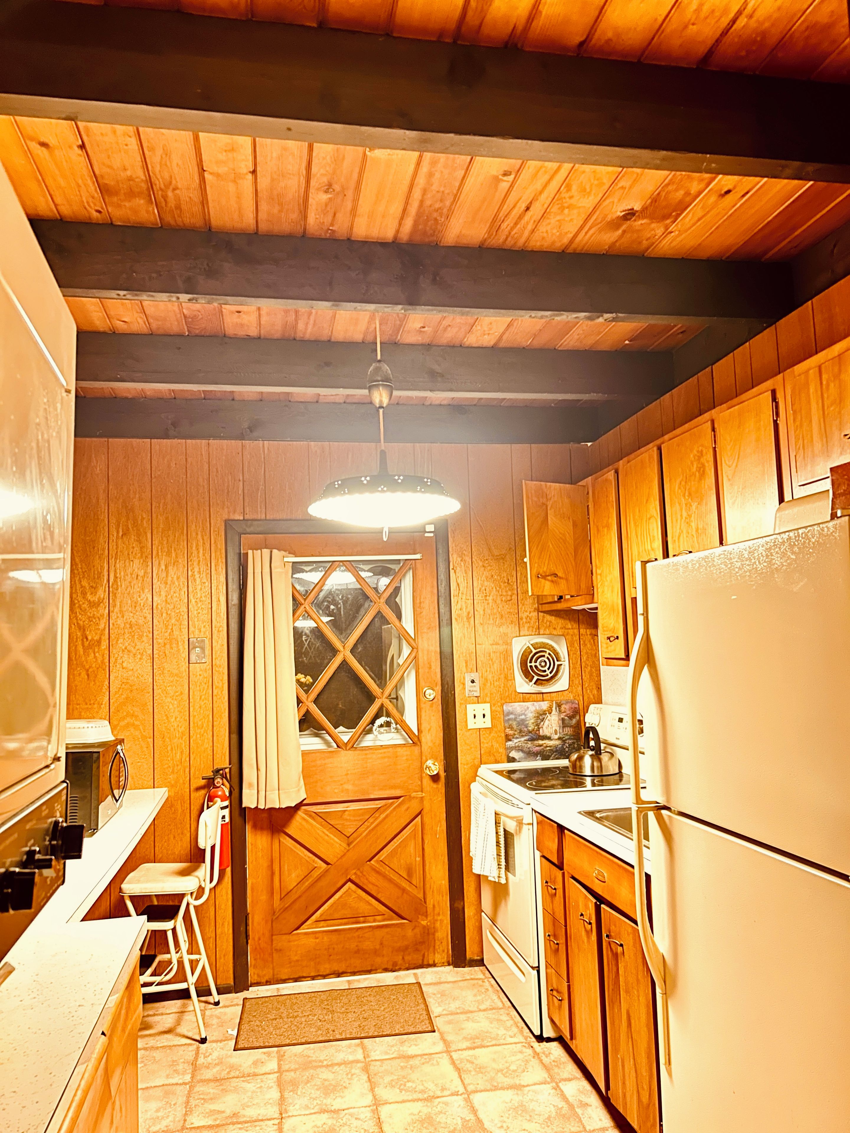 A compact kitchen with wooden cabinets, a refrigerator, a stove, and a hanging light fixture.