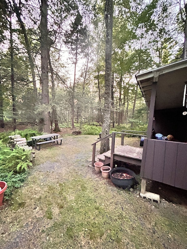 A forested area with a wooden deck, picnic table, and a barbecue grill.
