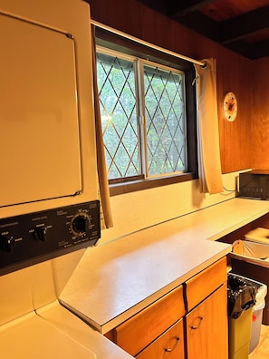 A compact kitchen with a washing machine, a window with a view of greenery, and a wooden cabinet.