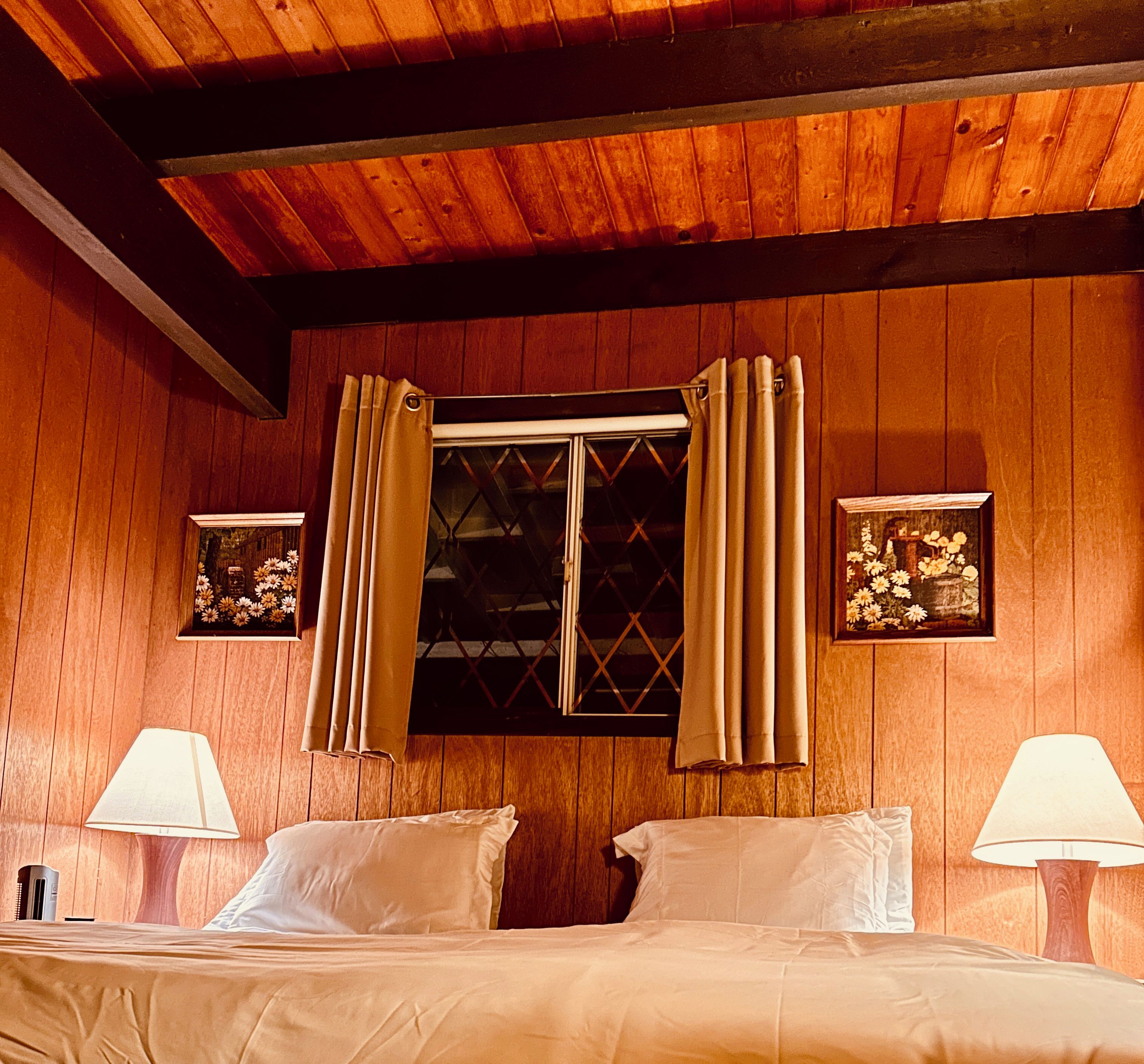 A wooden-paneled room with a bed, two bedside lamps, a window with curtains, and two framed pictures on the walls.