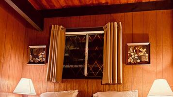 A wooden-paneled room with a bed, two bedside lamps, a window with curtains, and two framed pictures on the walls.