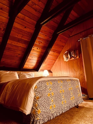 A bedroom with a wooden ceiling, a bed with a floral bedspread, and a nightstand with two hats.