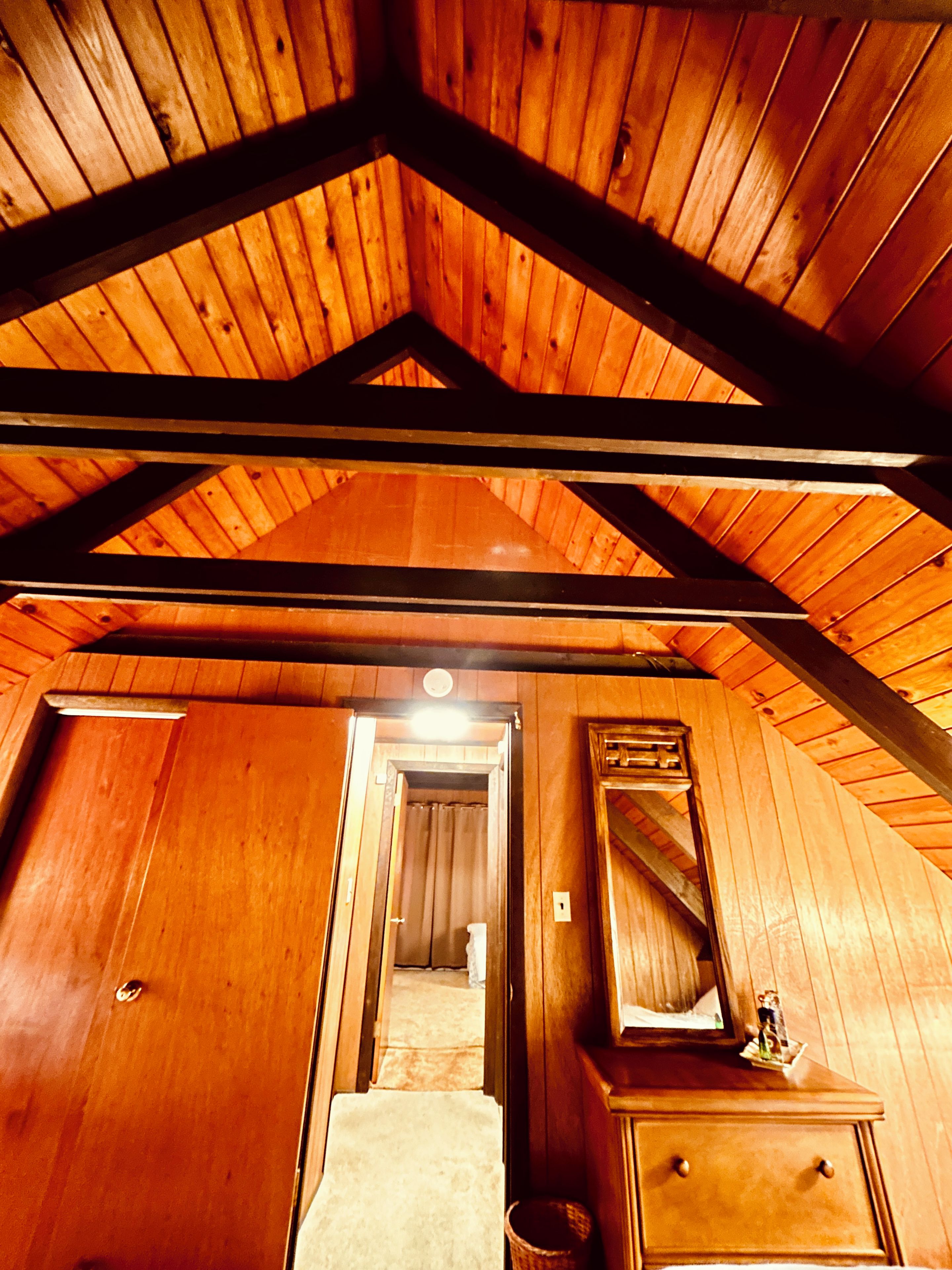A wooden interior with a sloped ceiling, a hallway leading to a bathroom, and a dresser with a mirror.