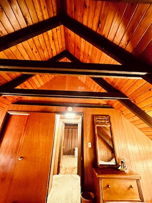 A wooden interior with a sloped ceiling, a hallway leading to a bathroom, and a dresser with a mirror.