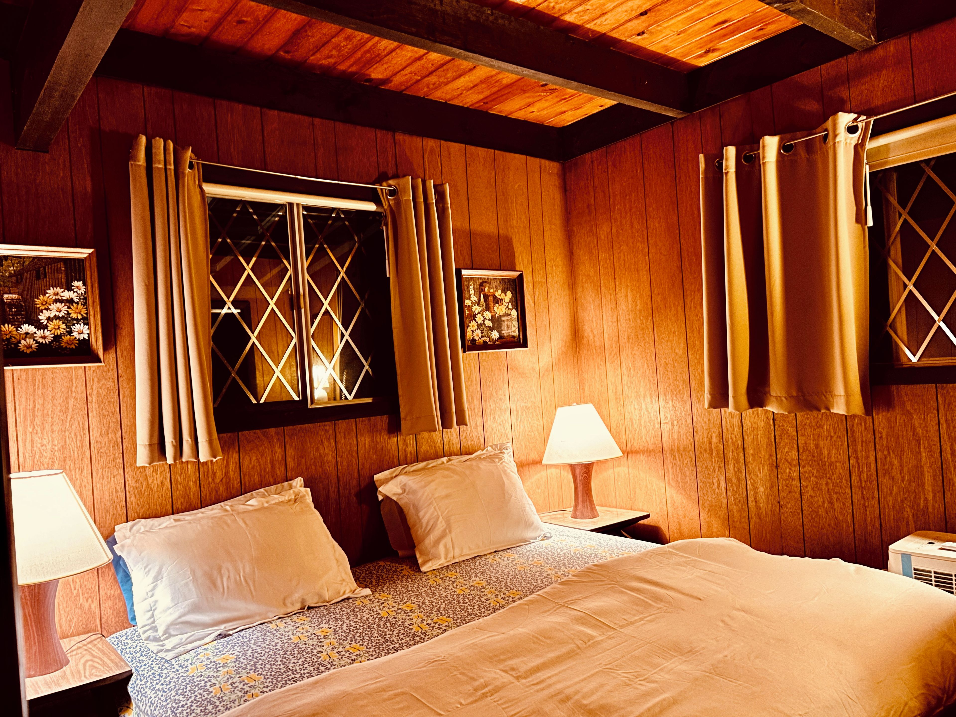 A room with wooden walls, a bed with white bedding, two bedside lamps, and a window with curtains.
