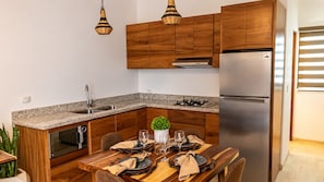 A modern kitchen with wooden cabinets, a stainless steel refrigerator, a granite countertop, and a dining area set with a table, chairs, and a potted plant. - Ancla Baja Living 4 (La Paz)