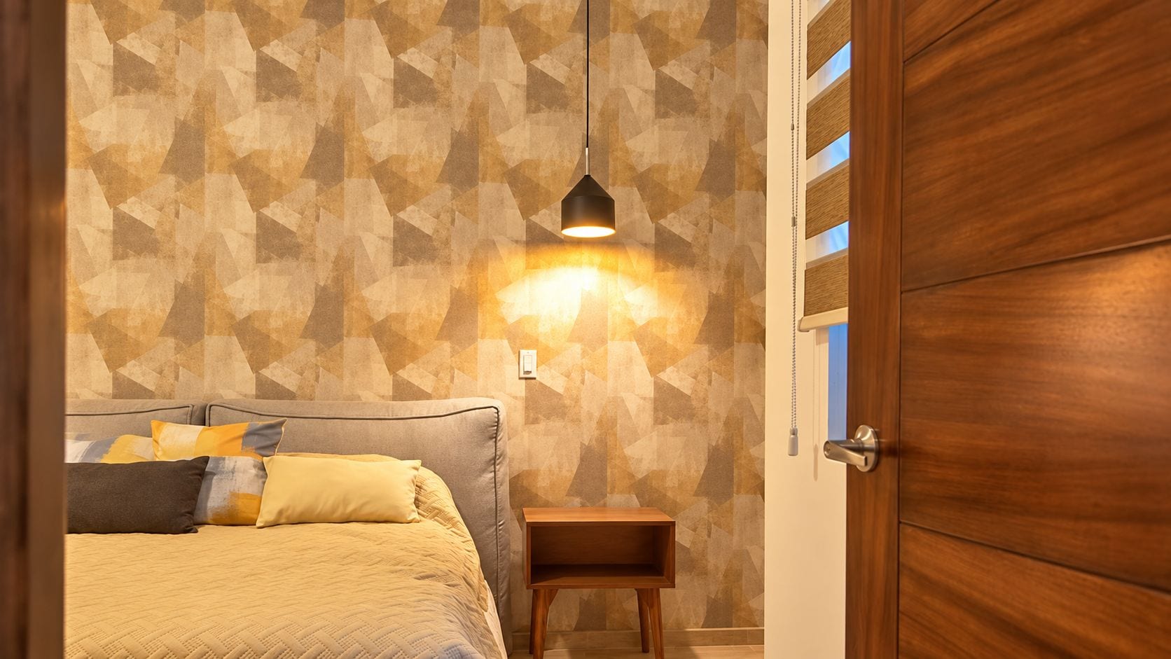 Une chambre à coucher avec un lit, des oreillers, une table de chevet, une lampe fixée au mur et une porte en bois.