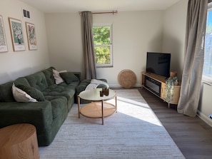 Een woonkamer met een hoekbank, een ronde salontafel, een televisie en een groot raam met gordijnen.
