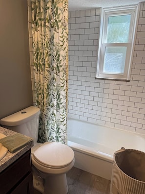 A bathroom with a toilet, a shower curtain with a leaf pattern, a bathtub with a white tiled wall, and a window above the tub. - Newly Furnished and Updated Apartment, Peaceful Neighbourhood (Euclid)