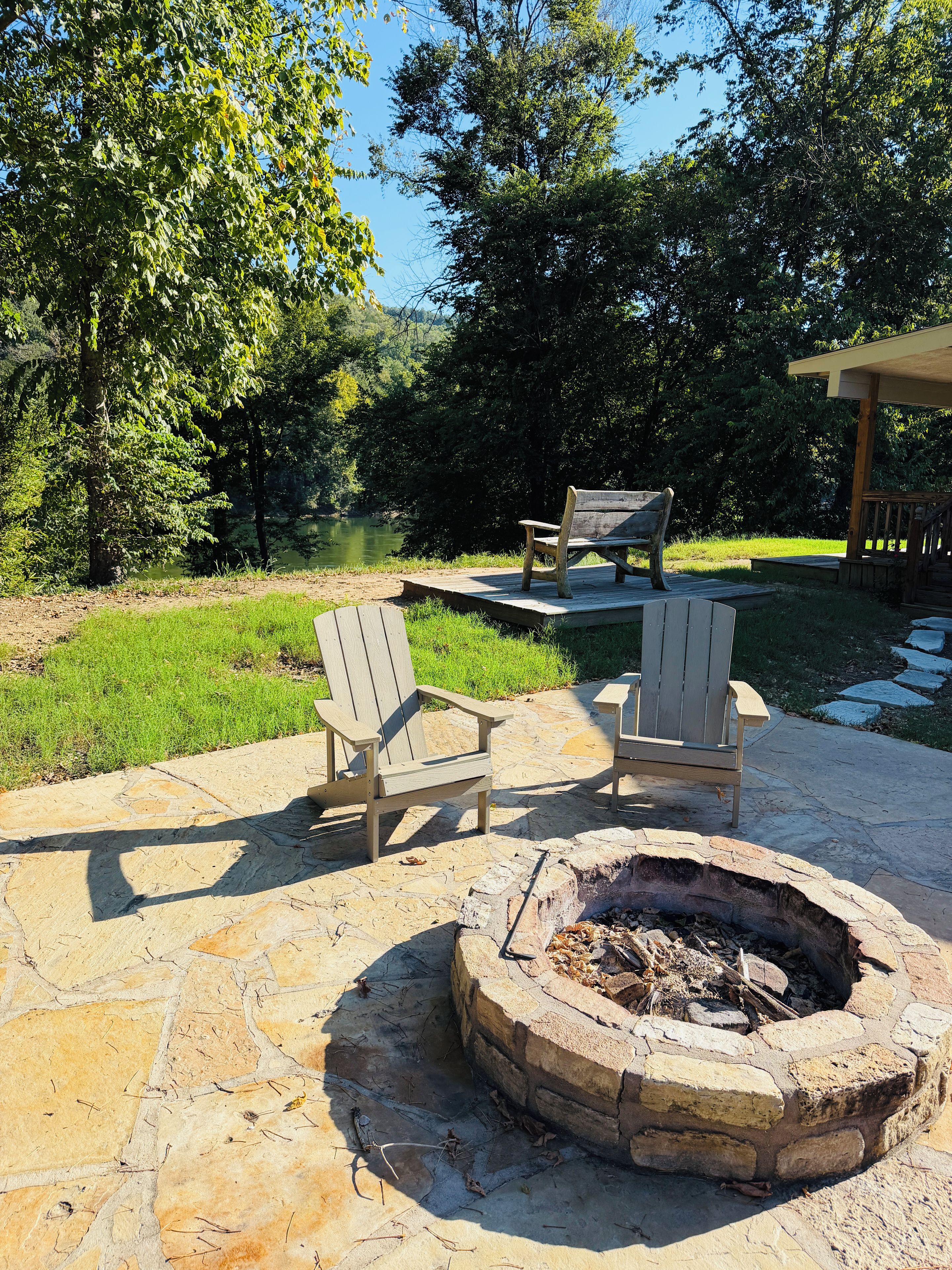An outdoor patio area with a stone fire pit, two Adirondack chairs, and a wooden deck overlooking a grassy area and trees.