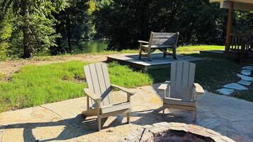An outdoor patio area with a stone fire pit, two Adirondack chairs, and a wooden deck overlooking a grassy area and trees.