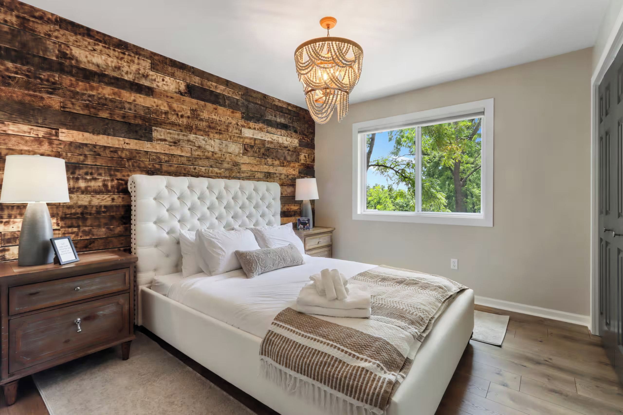 A bedroom with a wooden wall, a tufted headboard, a large window with a view of trees, and a chandelier.