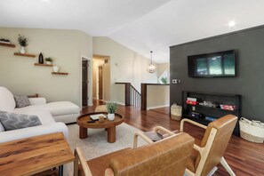 A modern living room with a sofa, coffee table, and chairs, a flat-screen TV mounted on a dark wall, and a staircase leading to an upper level. - Hillside Haven | Beach Access, Game Room & Fire Pit (Lake Geneva)
