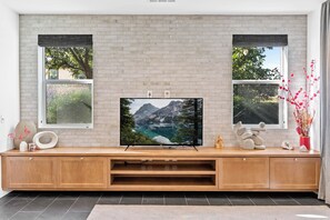 A modern living room with a large flat-screen TV mounted on a brick wall, wooden entertainment unit, and two windows with black blinds. - 4b3b modern house in Ontario/Model Home/Two Living room/AC/WIFI/back yard (Ontario)