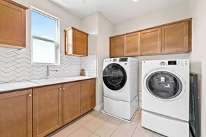 A laundry room with two front-loading washing machines, wooden cabinets, a window, and a tiled floor. - 4b3b modern house in Ontario/Model Home/Two Living room/AC/WIFI/back yard (Ontario)