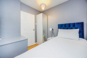 Een slaapkamer met een bed, nachtkastje, lamp en een deur.