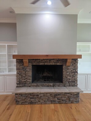 A stone fireplace with a wooden mantel in a room with wooden flooring and white walls.