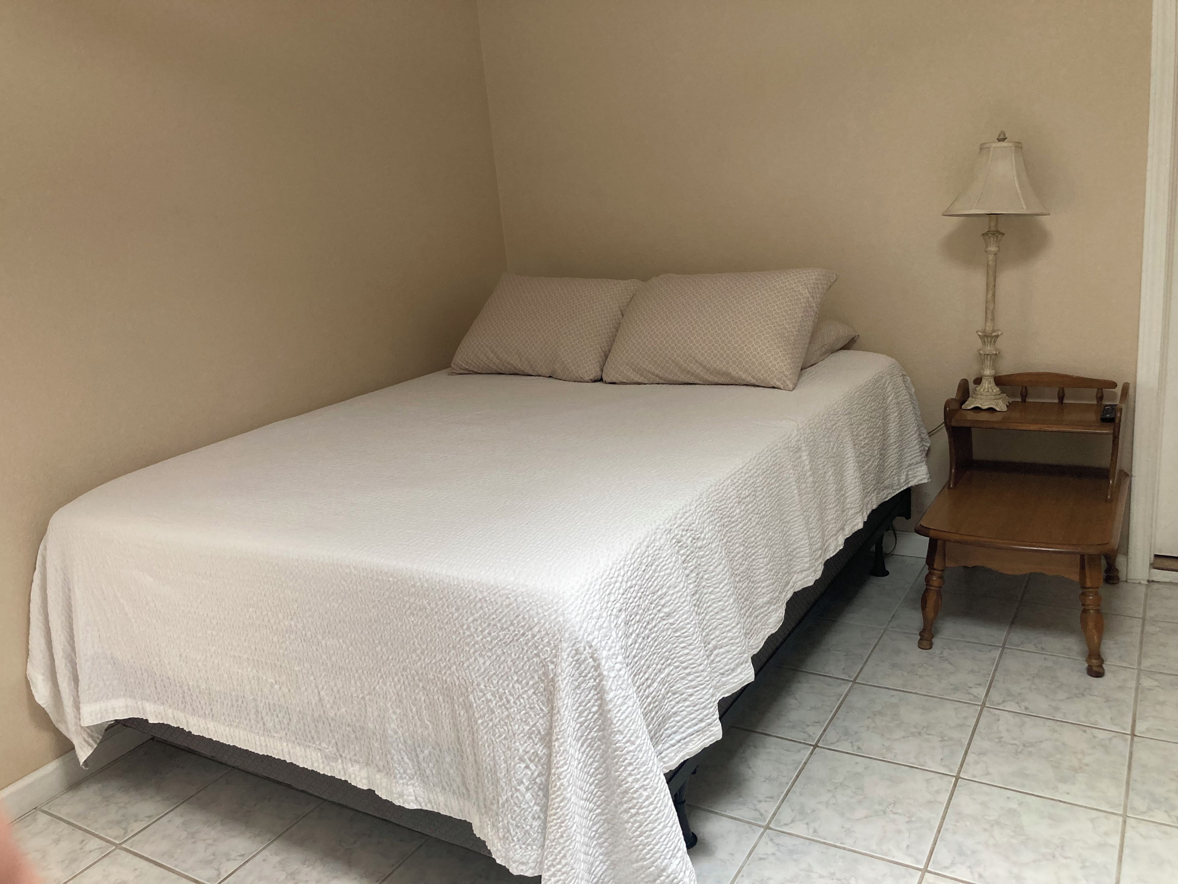Un letto singolo con copriletto bianco e due cuscini in una stanza d’angolo, con un piccolo comodino in legno e una lampada.