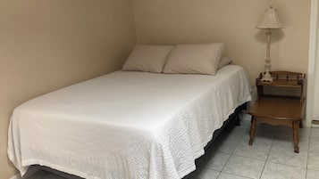 Un letto singolo con copriletto bianco e due cuscini in una stanza d’angolo, con un piccolo comodino in legno e una lampada.
