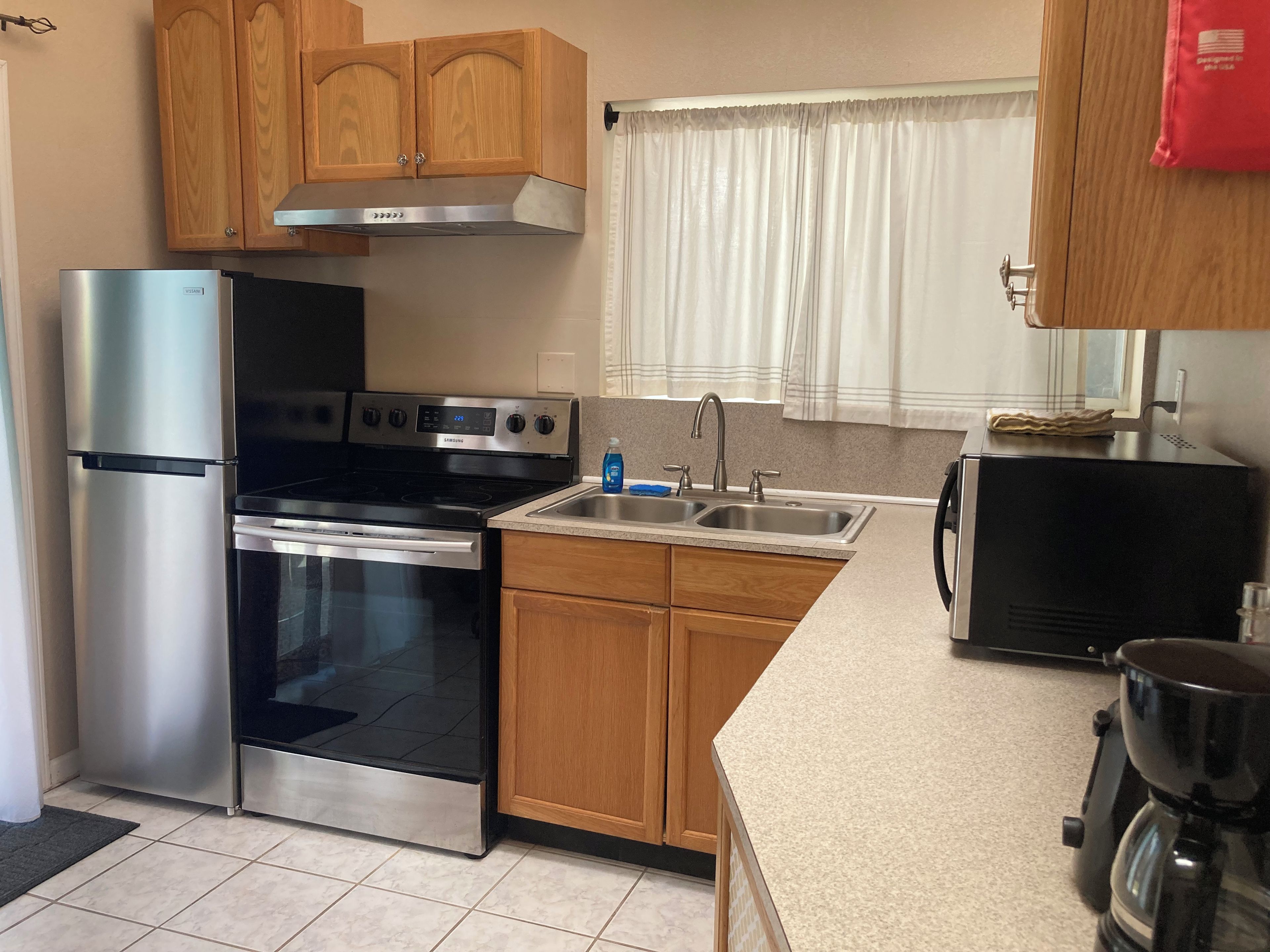 A kitchen with stainless steel appliances, wooden cabinets, a double sink, and a window with curtains.