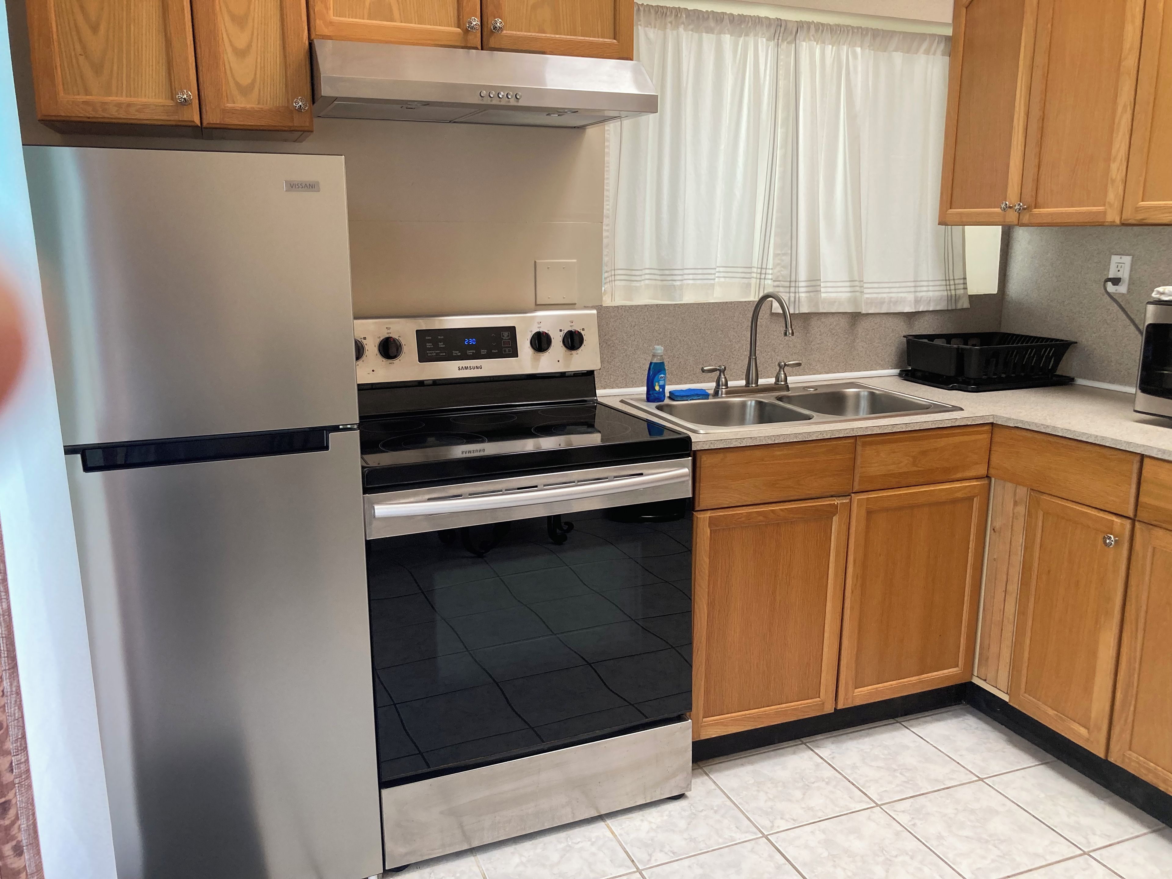 A kitchen with a stainless steel refrigerator, a built-in oven, a sink, wooden cabinets, and a window with curtains.