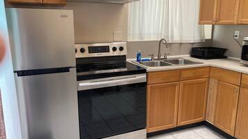 A kitchen with a stainless steel refrigerator, a built-in oven, a sink, wooden cabinets, and a window with curtains.