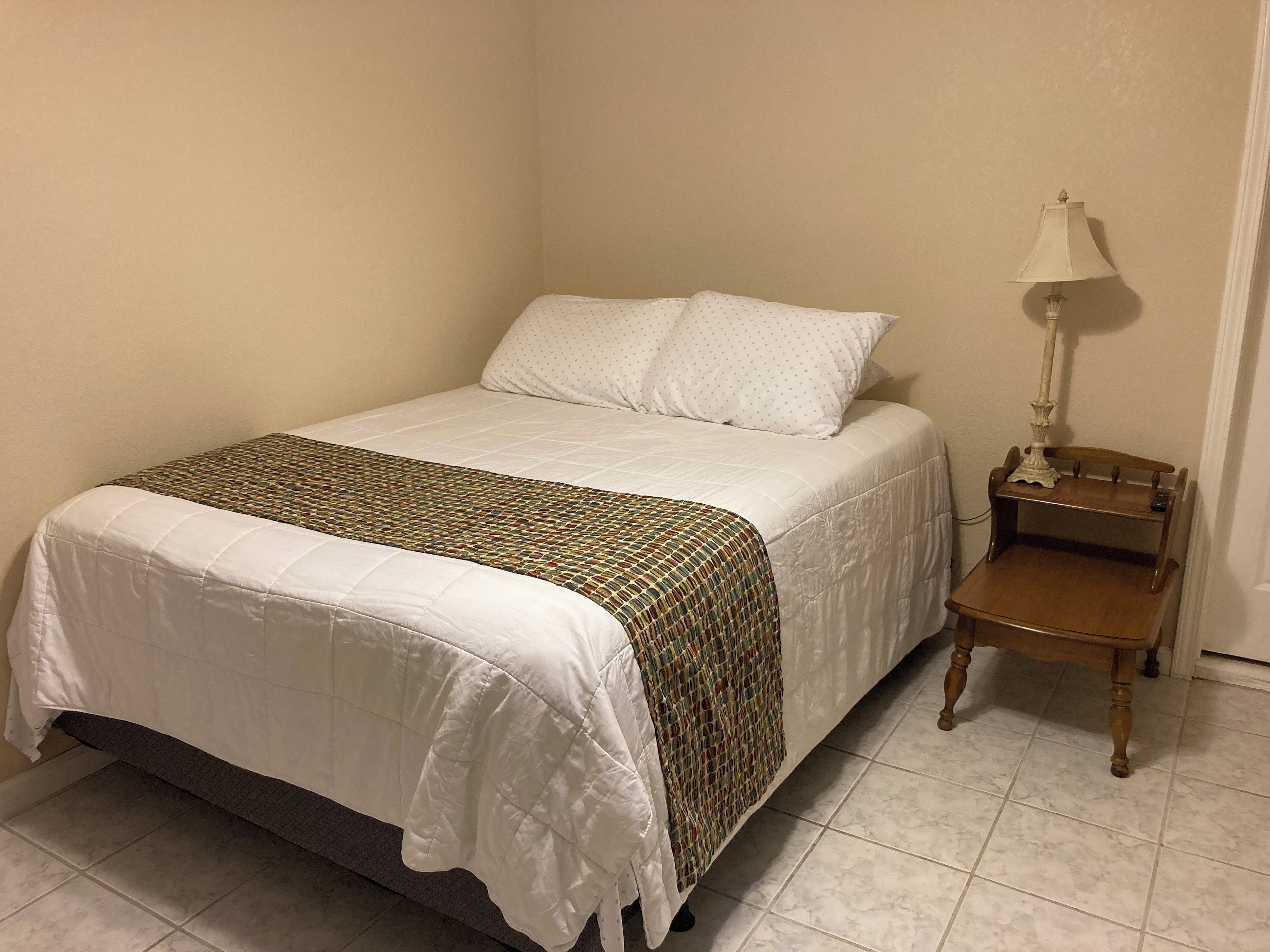 Een netjes opgemaakt bed met een gestreept beddekleed, een nachtkastje met een lamp en een stoel.