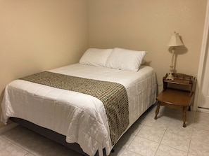Een netjes opgemaakt bed met een gestreept beddekleed, een nachtkastje met een lamp en een stoel.