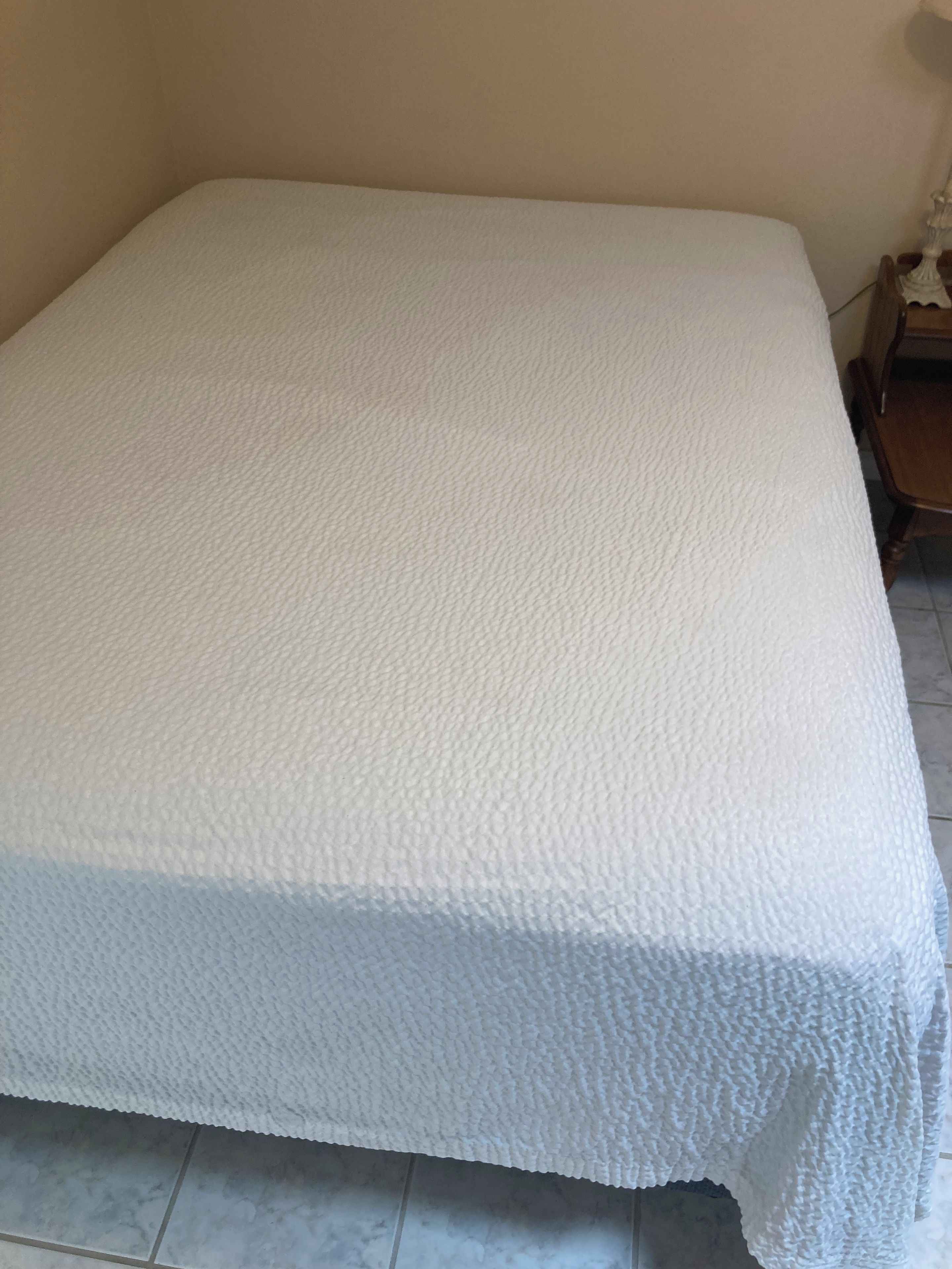Een bed met een beklede witte deken in een kamer met een betegelde vloer en een houten nachtkastje.