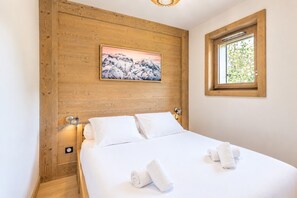 Ein Schlafzimmer mit einem hölzernen Kopfende, einem Gemälde von einer Berglandschaft, einem Fenster mit Blick auf Grünflächen und zwei aufgerollten Handtüchern auf dem Bett.