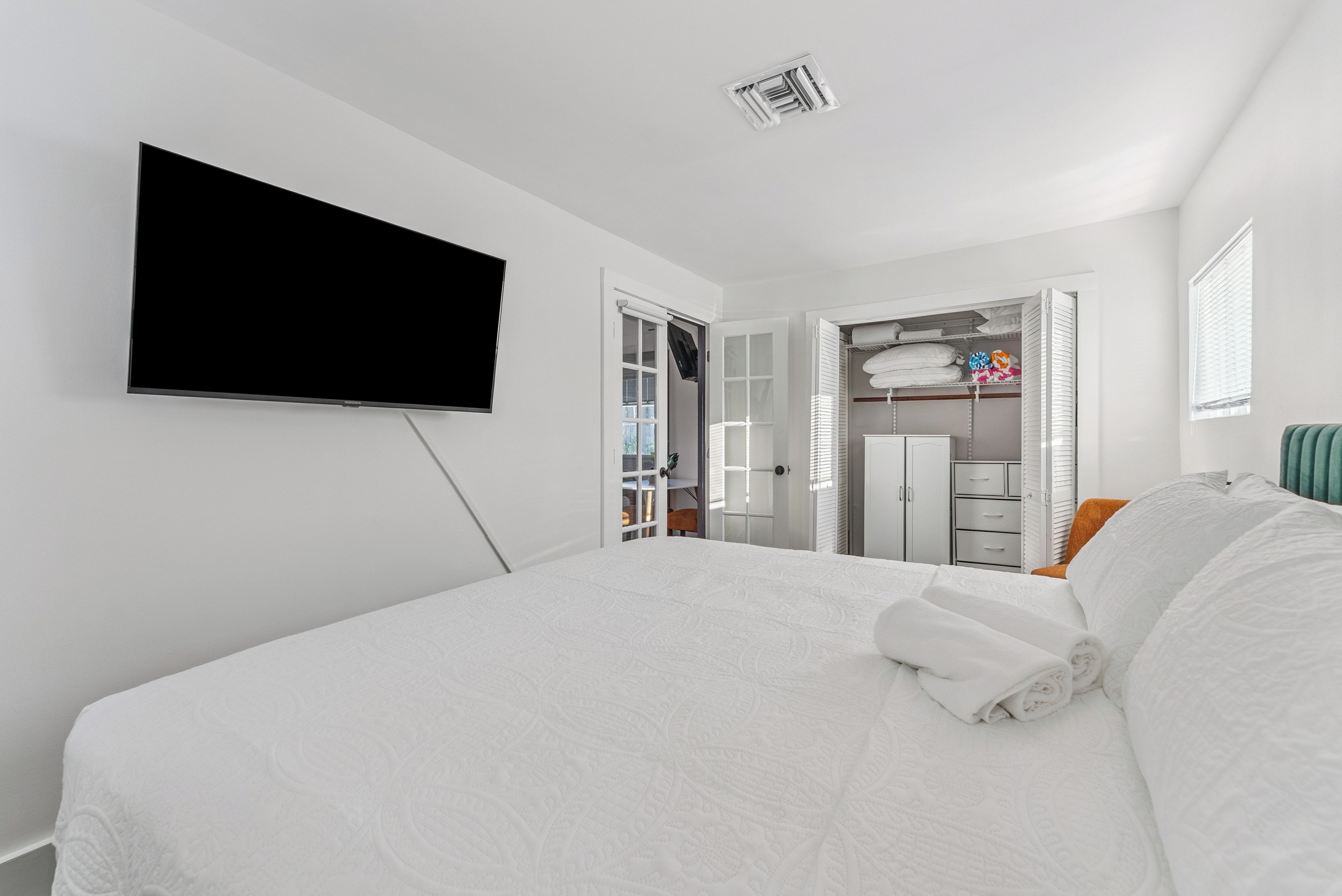 A hotel room with a bed, a flat-screen TV mounted on the wall, a walk-in closet, and a window with blinds.
