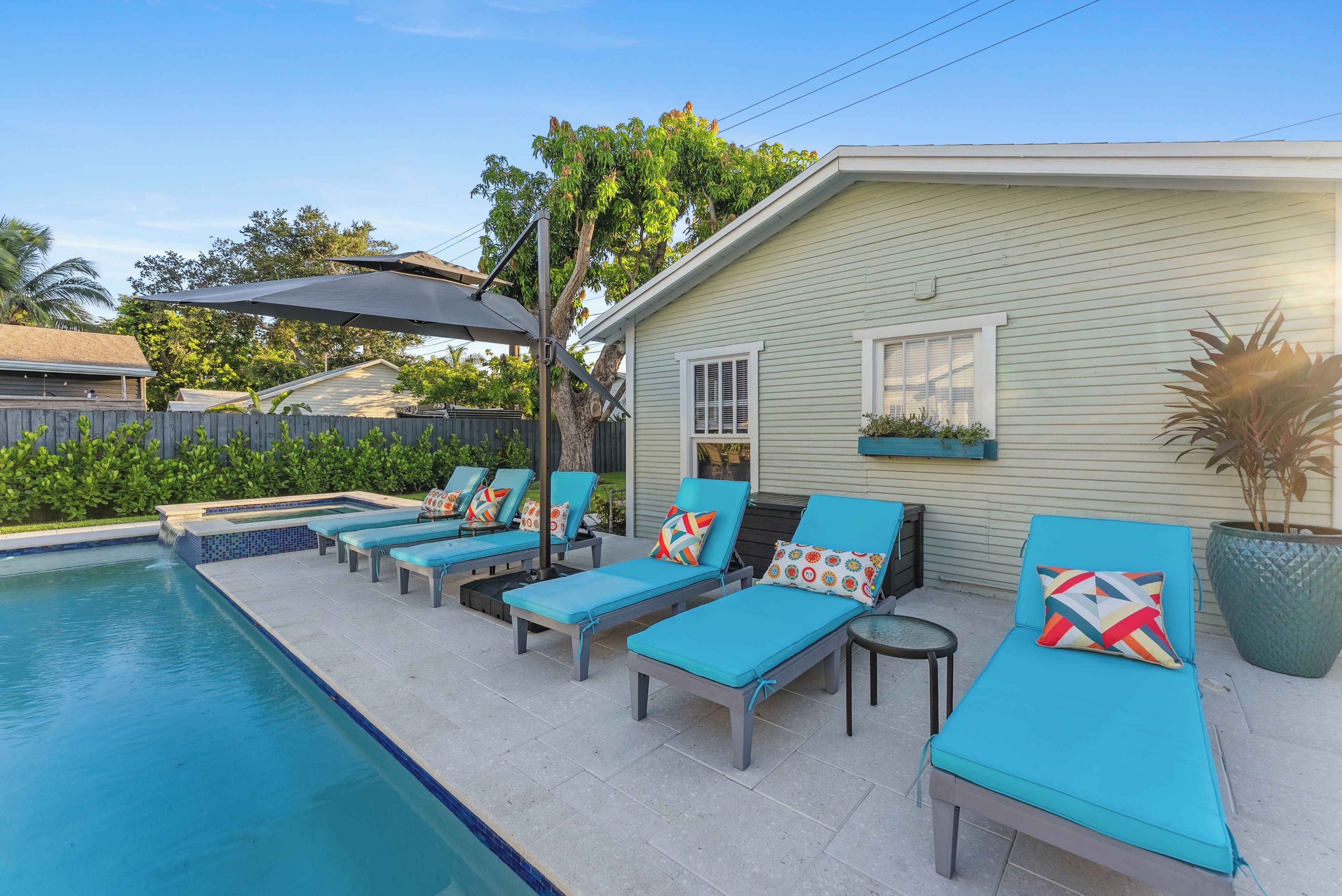 A poolside area with lounge chairs and a small table, a covered patio, and a house with a window and a planter.