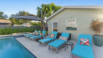 A poolside area with lounge chairs and a small table, a covered patio, and a house with a window and a planter.