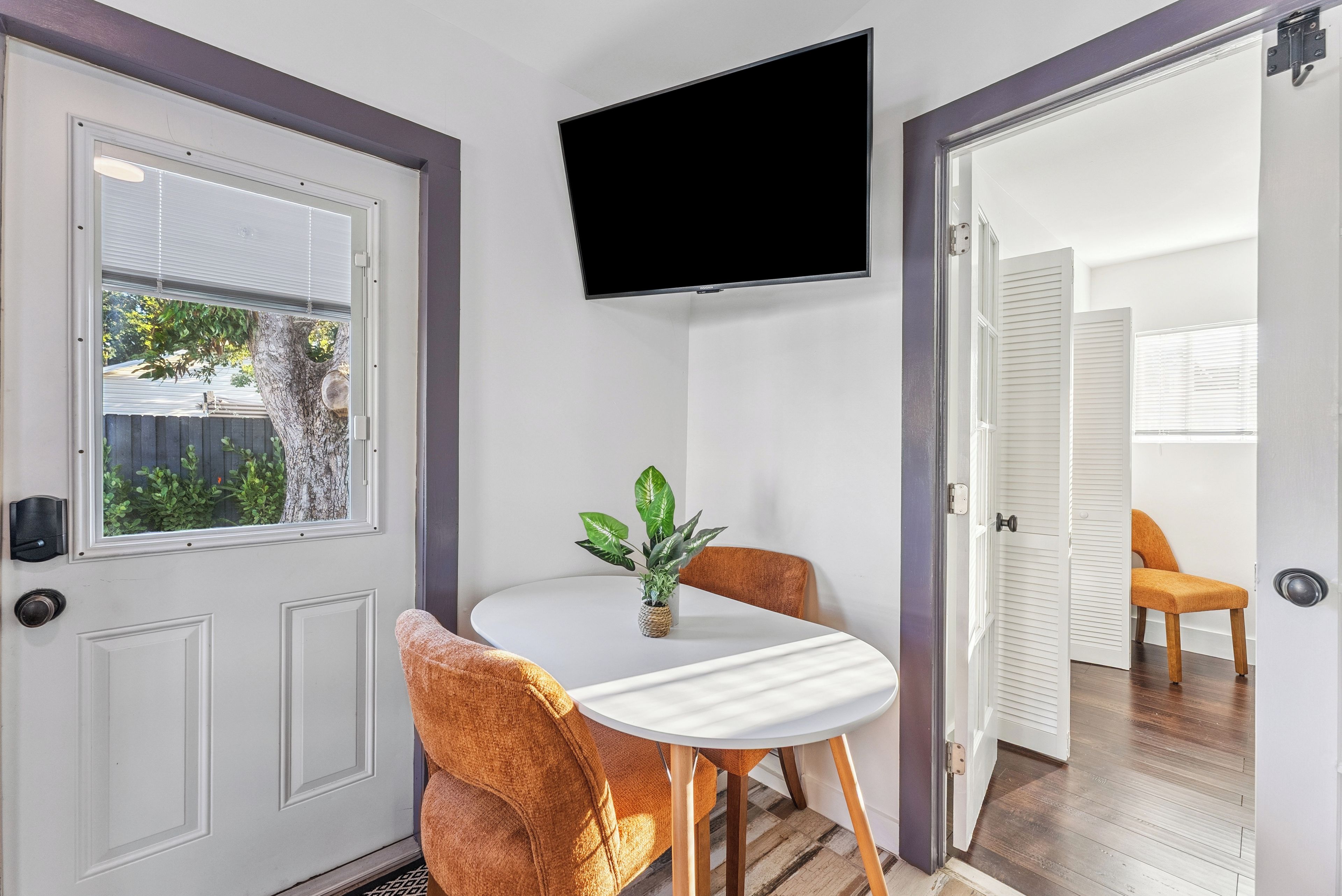 Ein Zimmer mit weißer Tür, einem an der Wand befestigten Fernseher, einem kleinen Tisch mit Stühlen und einem Blumentopf.