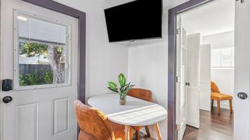 Ein Zimmer mit weißer Tür, einem an der Wand befestigten Fernseher, einem kleinen Tisch mit Stühlen und einem Blumentopf.