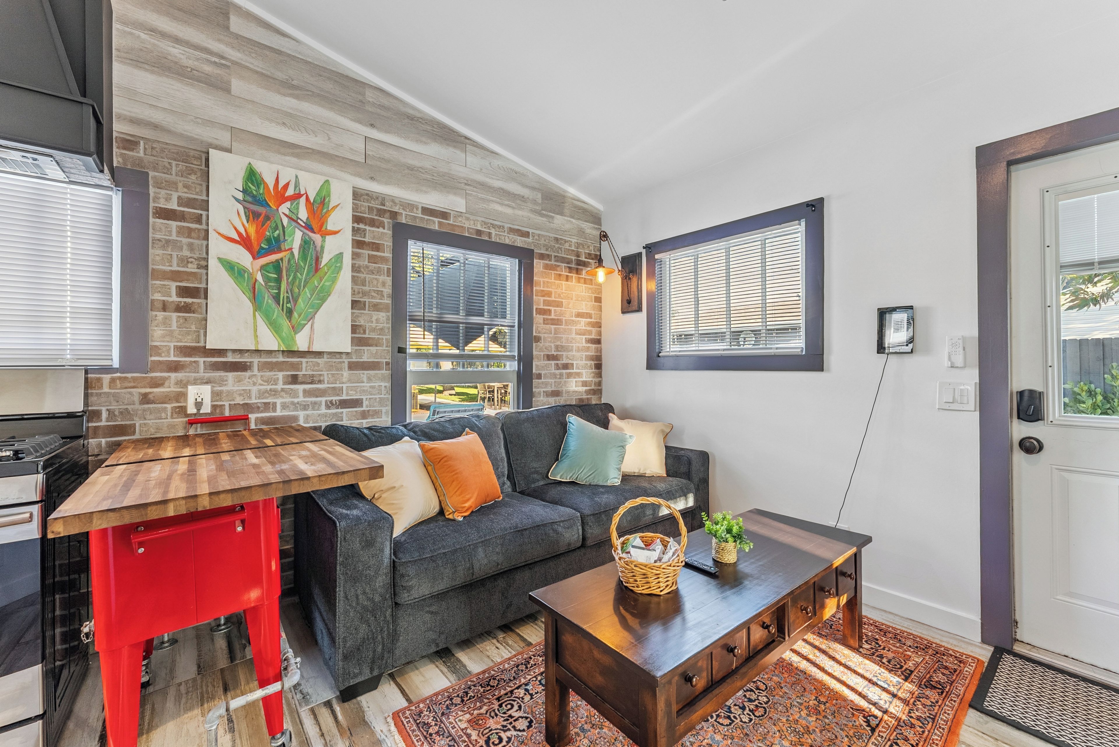 A modern living room with a brick wall, a wooden dining table, a grey sofa with cushions, and a red kitchen counter.