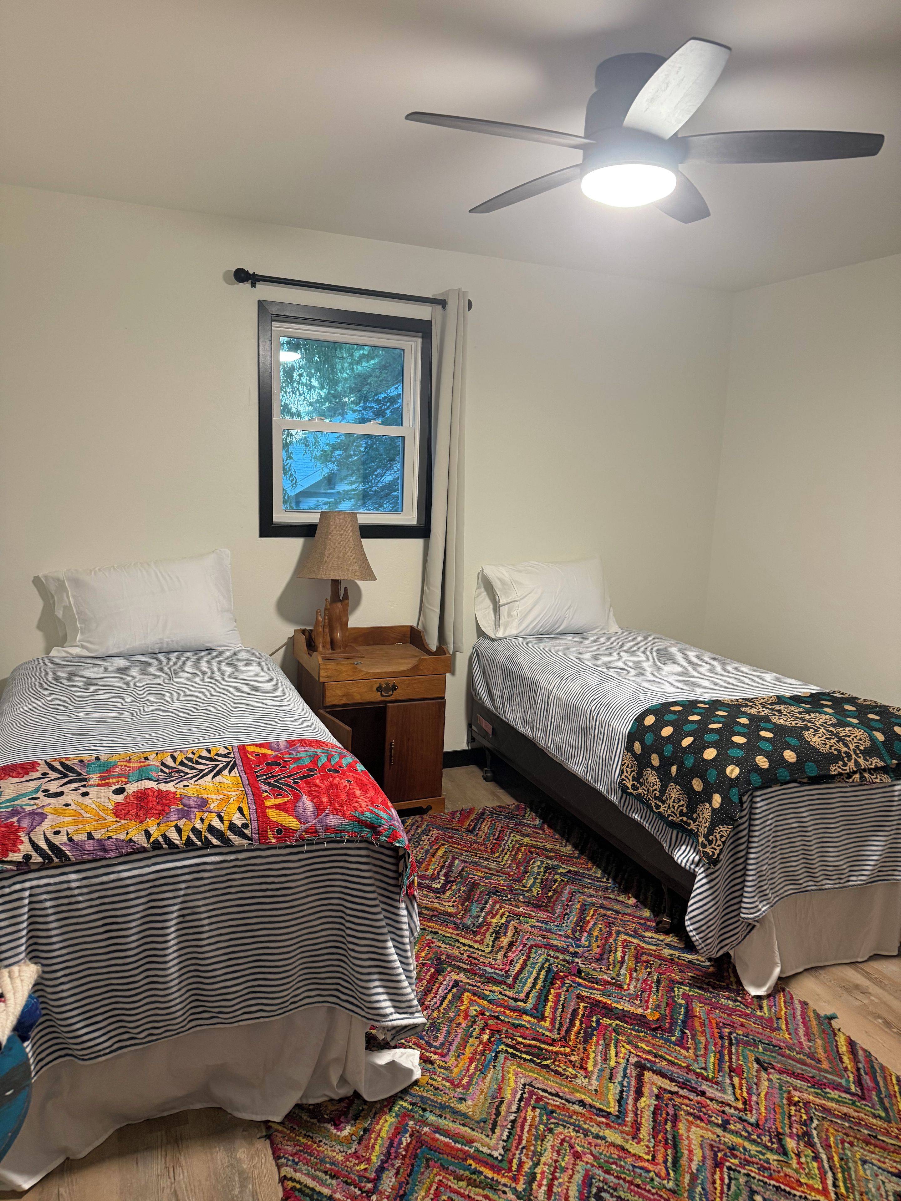 A bedroom with two beds, a ceiling fan, a window, and a colorful rug.