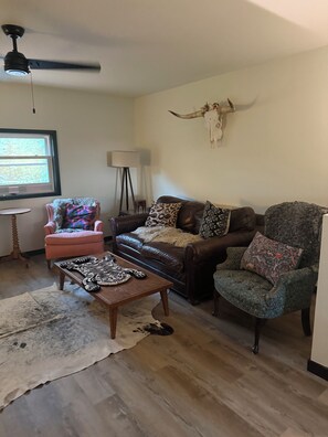 A living room with a brown leather sofa, a pink armchair, a patterned armchair, a coffee table with a zebra print, a cowhide rug, and a ceiling fan.