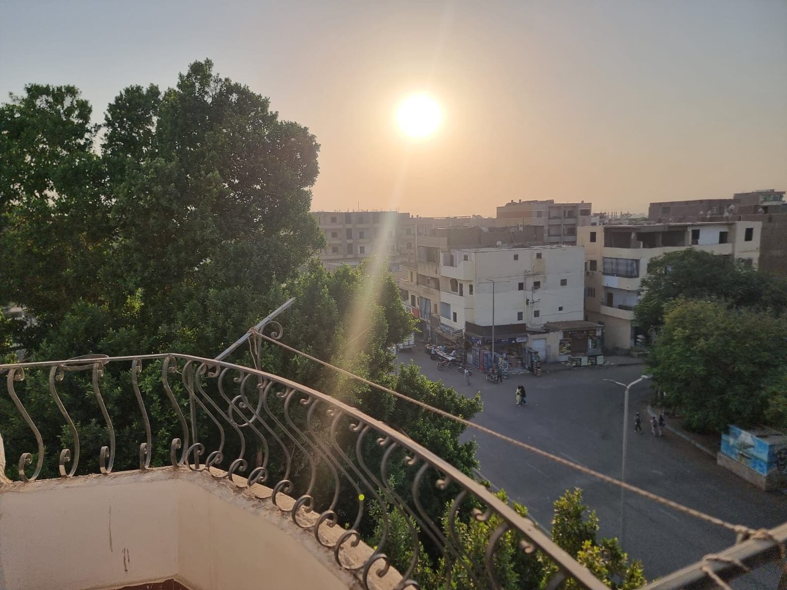 Un balcon doté d’une rambarde en métal, donnant sur une rue bordée d’immeubles et d’arbres.