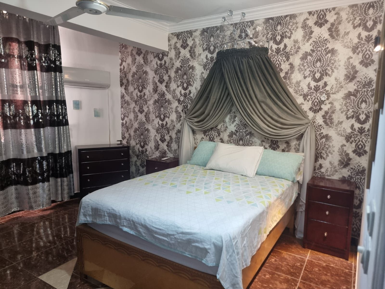 A bedroom with a bed, curtains, a dresser, and patterned wallpaper.