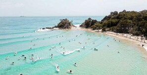 Ein Strand mit klarem türkisblauem Wasser, Menschen beim Schwimmen und Sonnenbaden sowie ein mit Vegetation bewachsener Felsen.