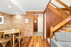 A room with a brick wall, wooden flooring, a dining table with chairs, a staircase with wooden railings, and a gray sofa. - Kipling Beachside Retreat (Byron)