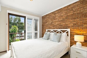 A bedroom with a brick wall, a bed with white bedding, a nightstand with a lamp, and a view of greenery through a sliding door. - Kipling Beachside Retreat (Byron)