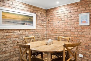 A room with a brick wall, a round wooden table with chairs, and two framed pictures. - Kipling Beachside Retreat (Byron)